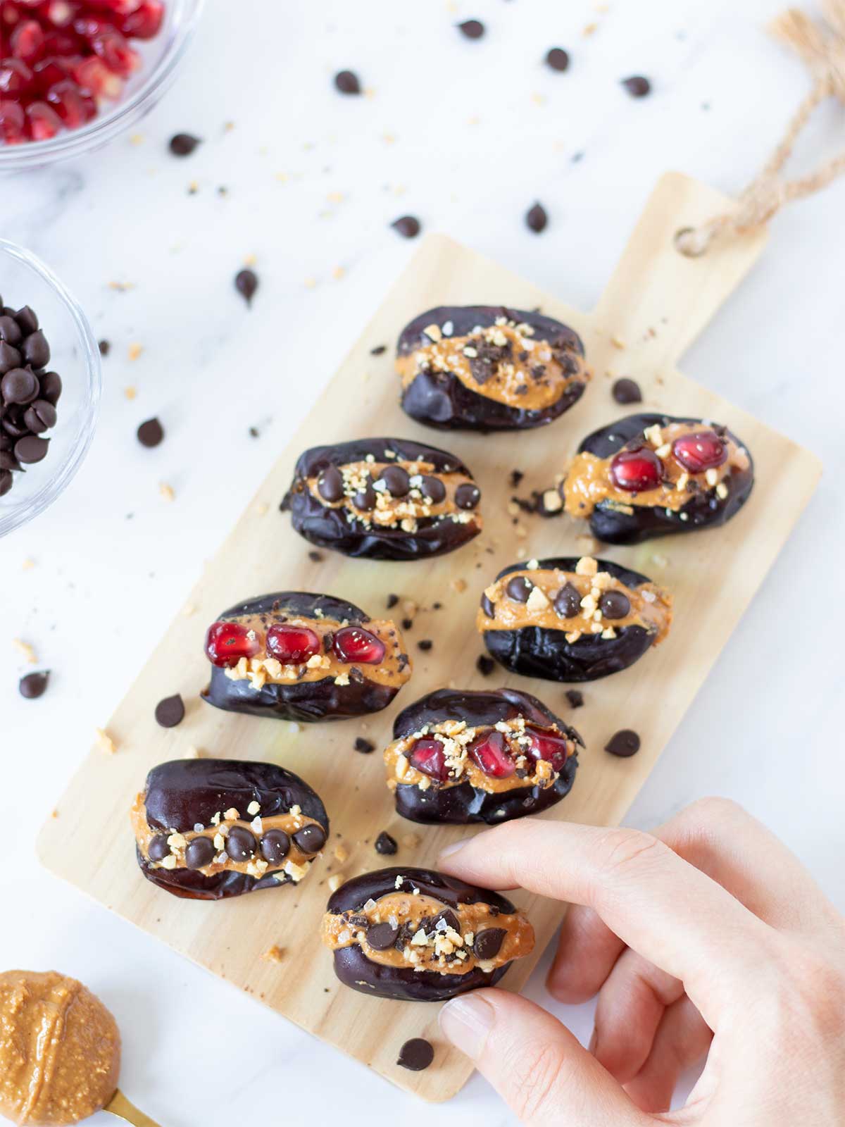 Soft and chewy dates stuffed with homemade peanut butter on small wooden board topped with chocolate chips, chopped dark chocolate, and pomegranate seeds