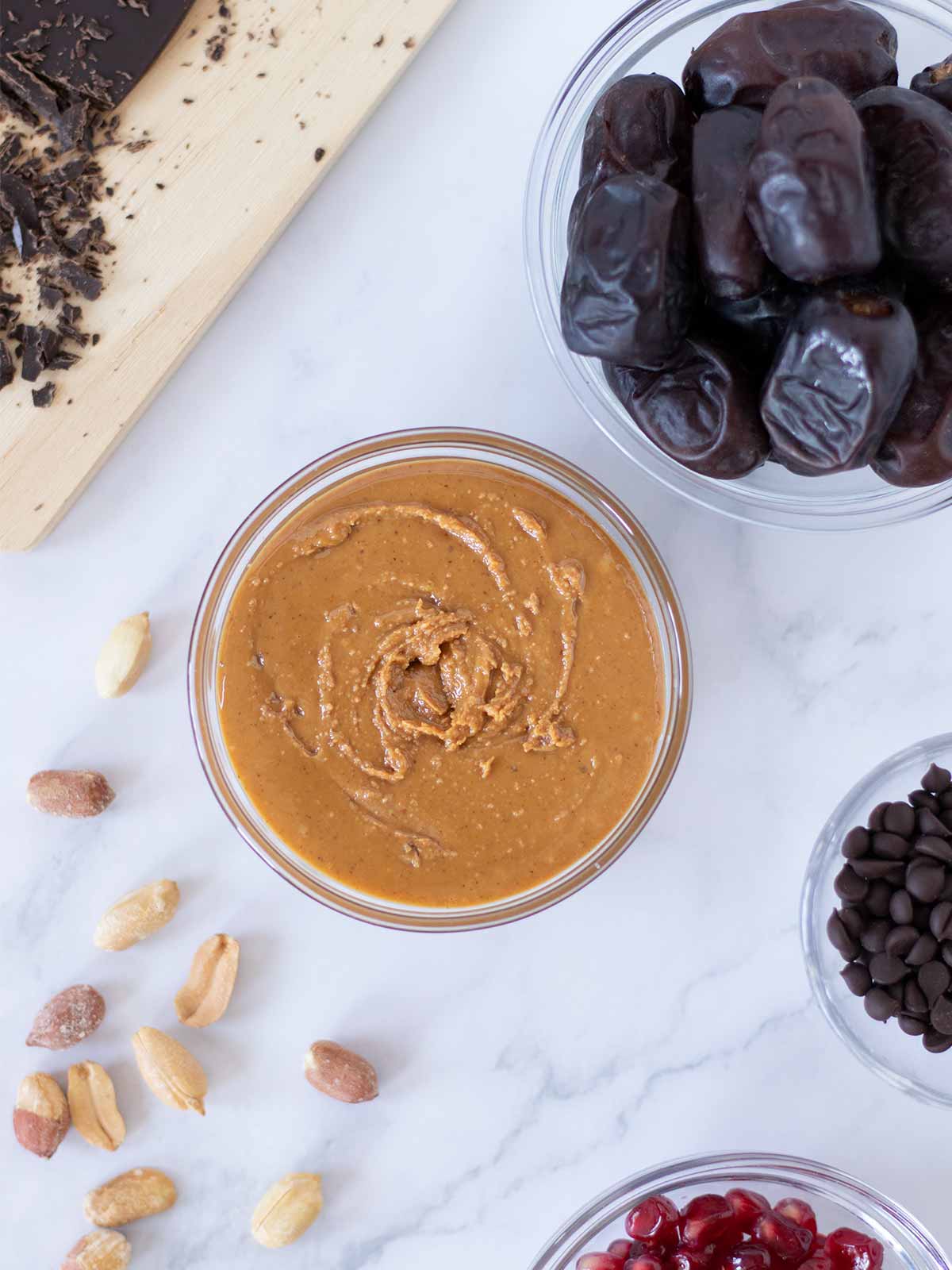 Simple plant-based ingredients for making refined sugar free dessert: Mazafati dates, creamy peanut butter, roasted peanuts, dark chocolate, chocolate chips, and fresh pomegranate arils