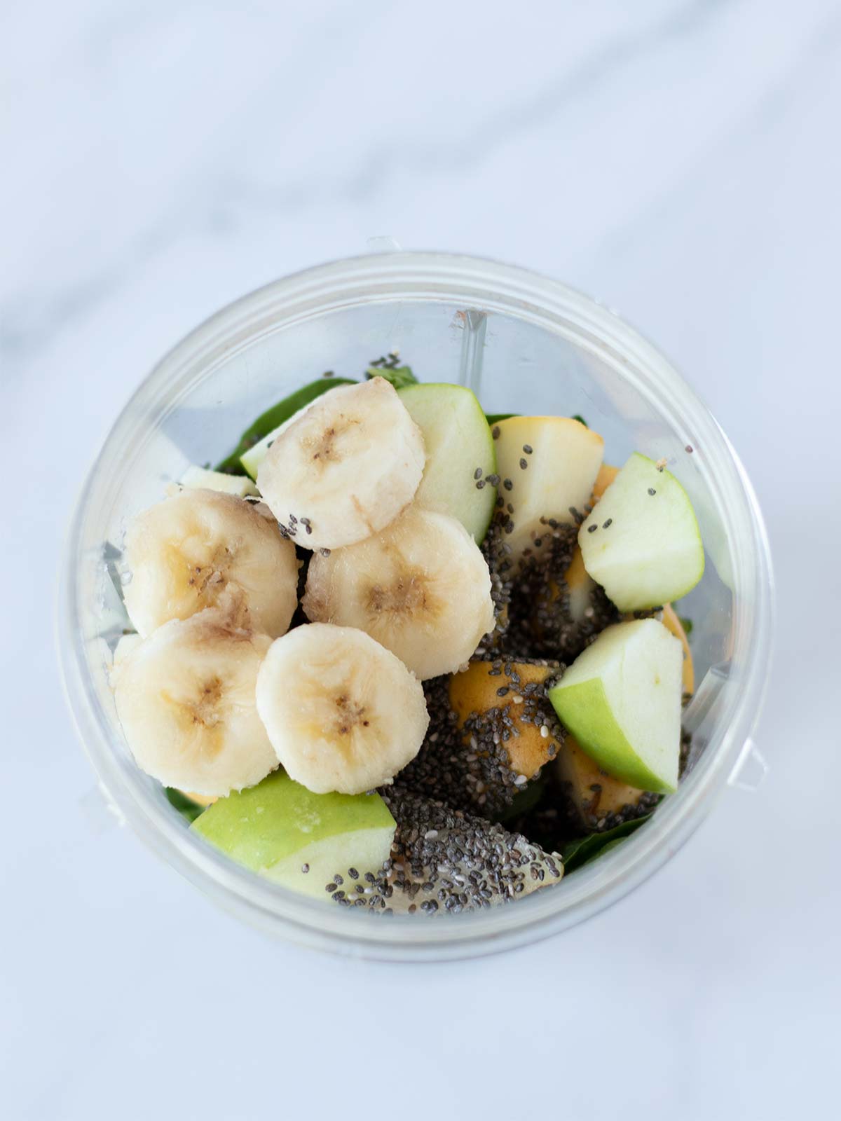 Chopped frozen banana, green apple, pear, chia seeds, lemon juice, spinach, and oat milk in a blender cup.