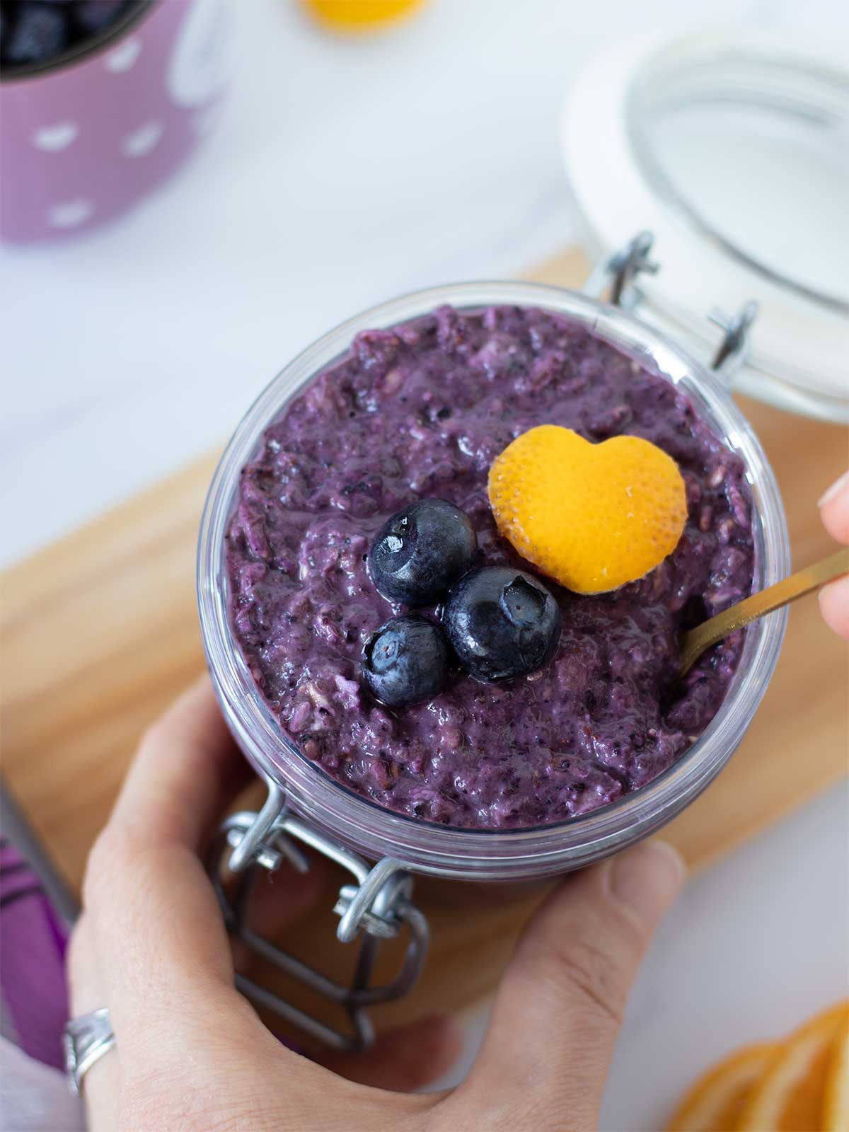 Hand holding creamy blueberry lemon overnight oats in a jar with golden spoon