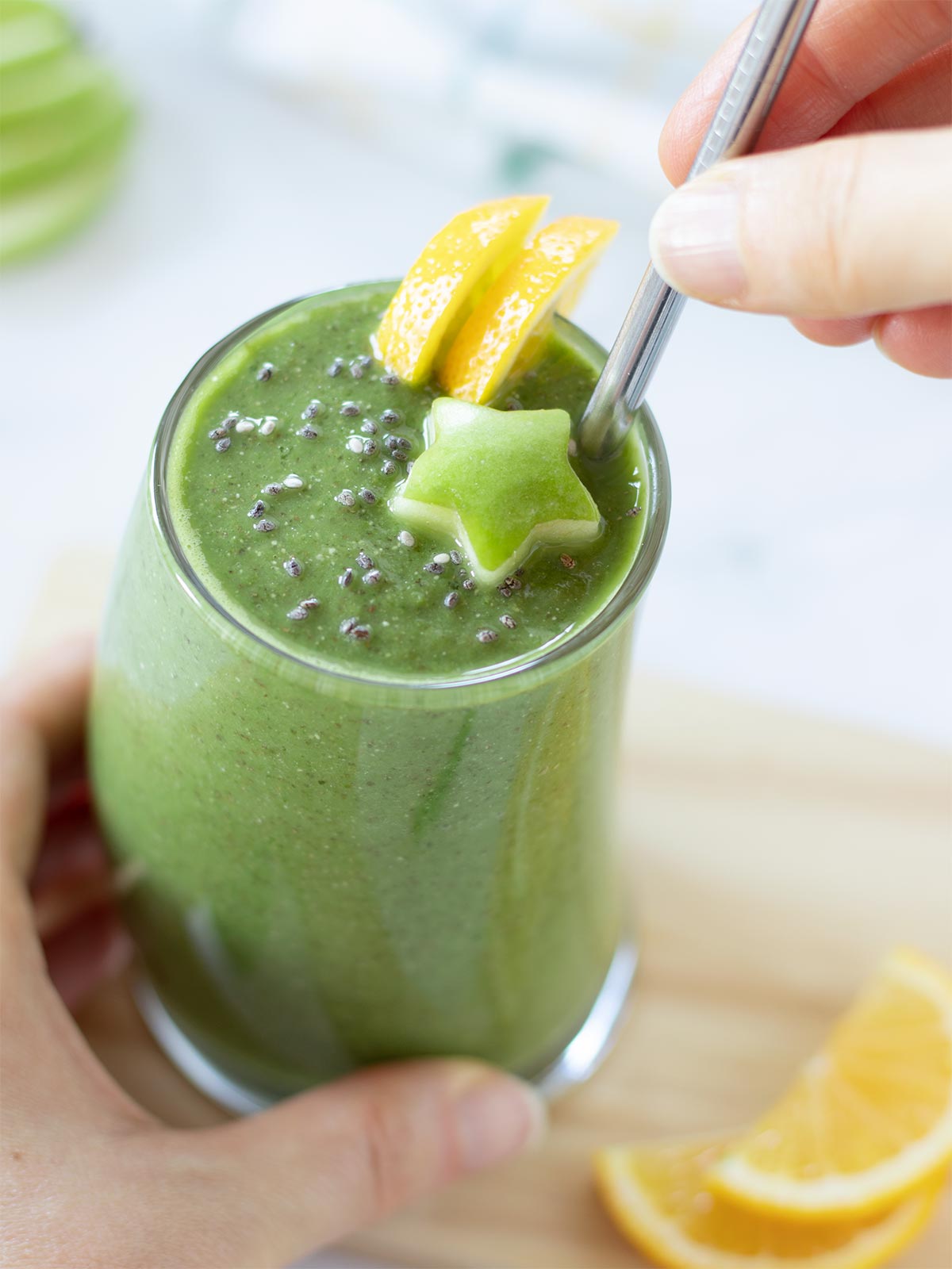 Female hand is holding a creamy homemade gut cleansing drink in a glass garnished with sliced lemon, chia seeds, and carved apple star with eco-friendly straw.
