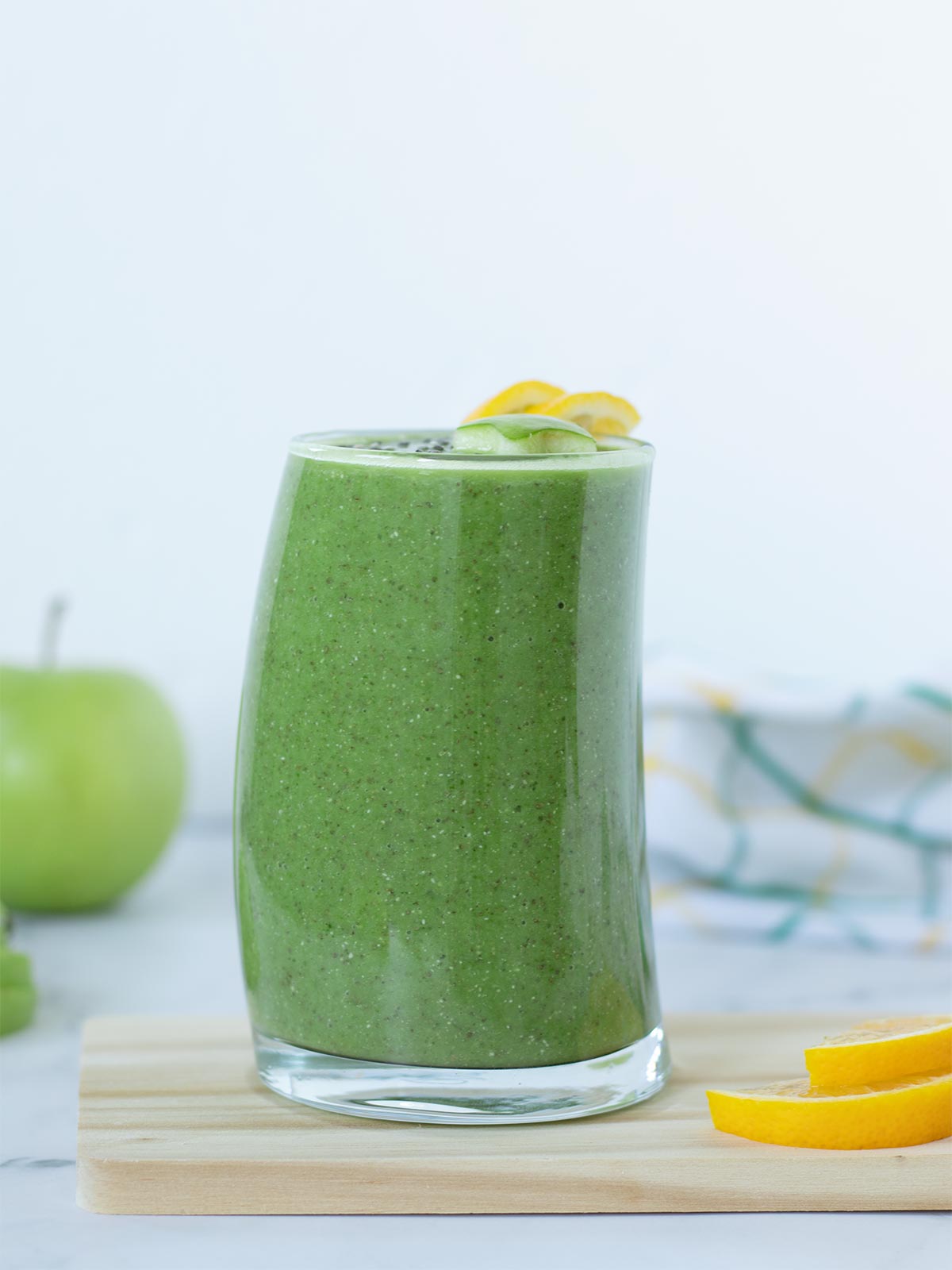 Refreshing green detox beverage in a tall glass on wooden boar with sliced lemon and green apple in the background.