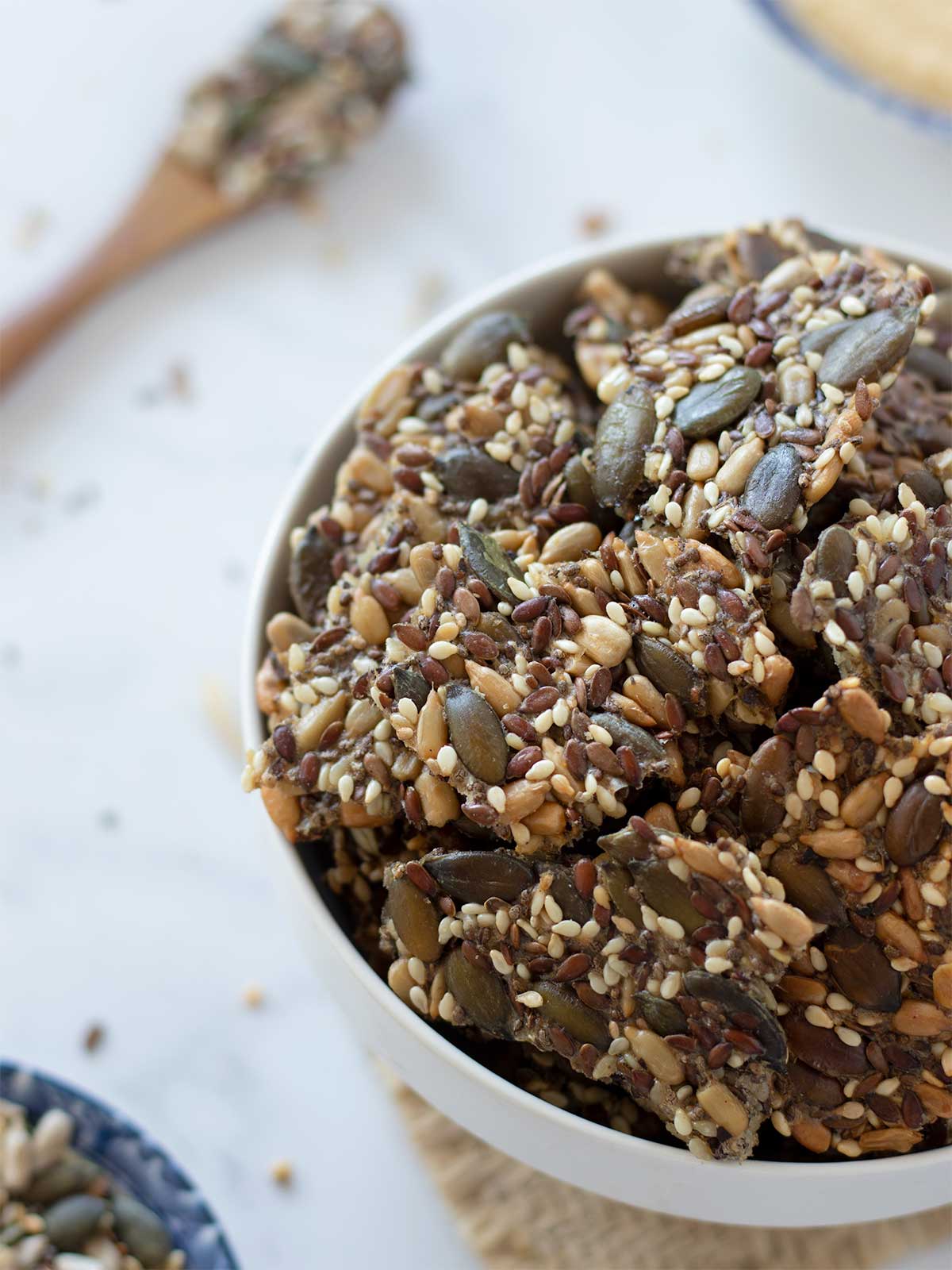 Crispy homemade super seed crackers without yeast or baking soda in a bowl on white table with wooden spoon and small plate filled with mixed seeds