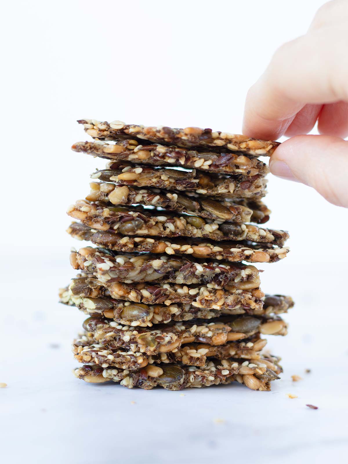 A stack of unleavened homemade healthy seed crackers on white with female hand holding the top piece of it