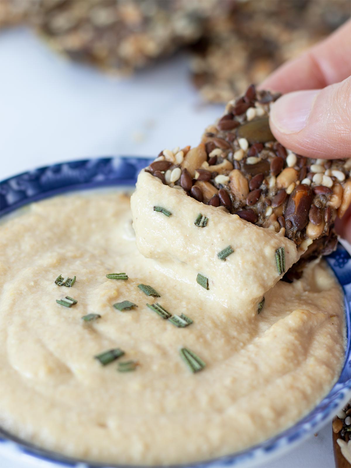 5 Super sseds crackers in female hand dipped in creamy homemade hummus garnished with chopped fresh rosemary