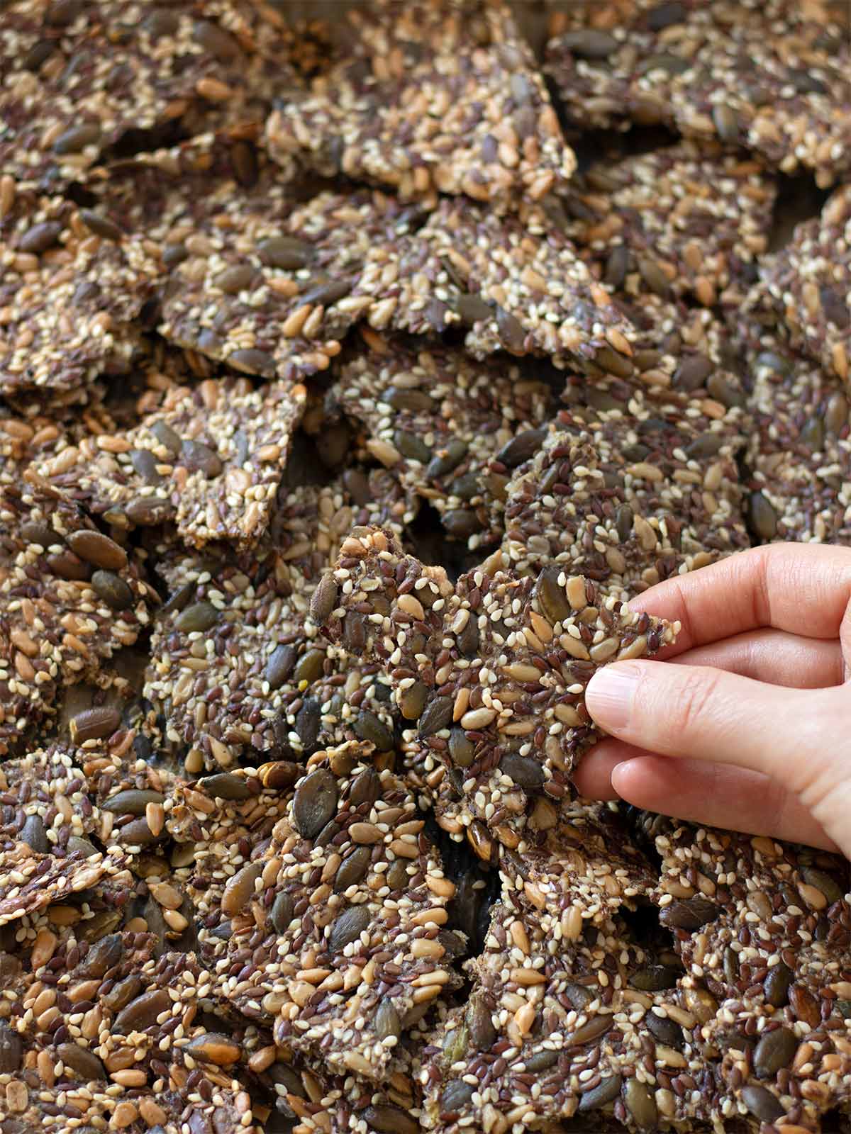Hand is holding healthy vegan and keto homemade savory cracker over a baking sheet full of crunchy freshly baked crackers