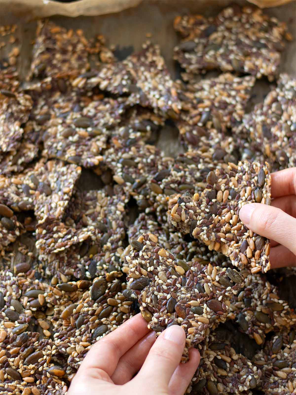Female hands breaking yeast-free seed crackers over a baking sheet full of healthy savory homemade crackers