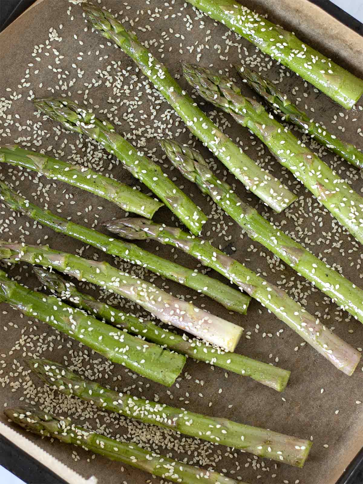 Fresh asparagus in baking sheet lined with parchment papper coated with extra virgin olive oil, salt and seasonings sprinkled with sesame seeds prepared for roasting in the oven