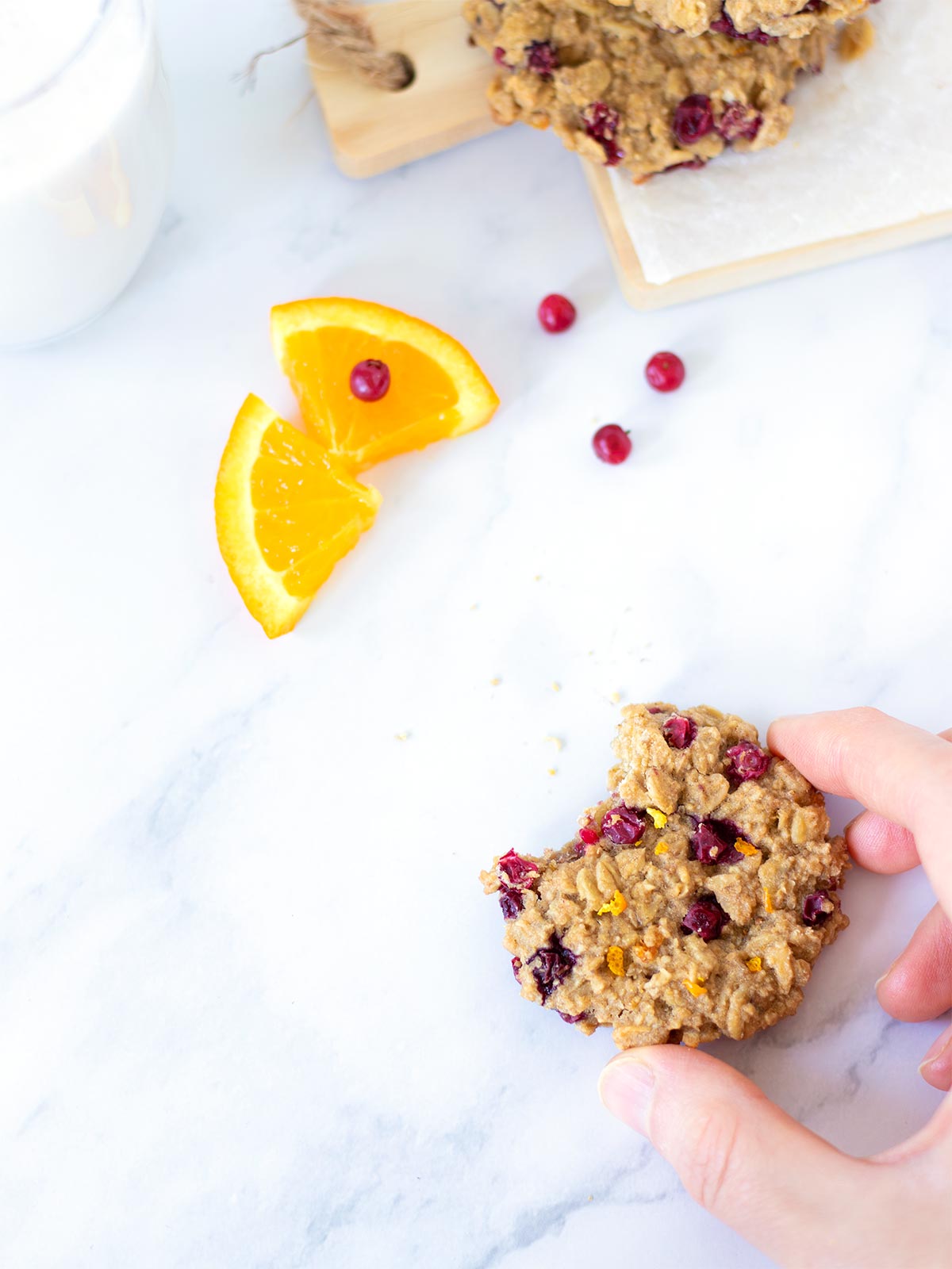 Female hand is holding vegan cranberry cookie with orange juice and zest on white marble table with a glass of milk for breakfast