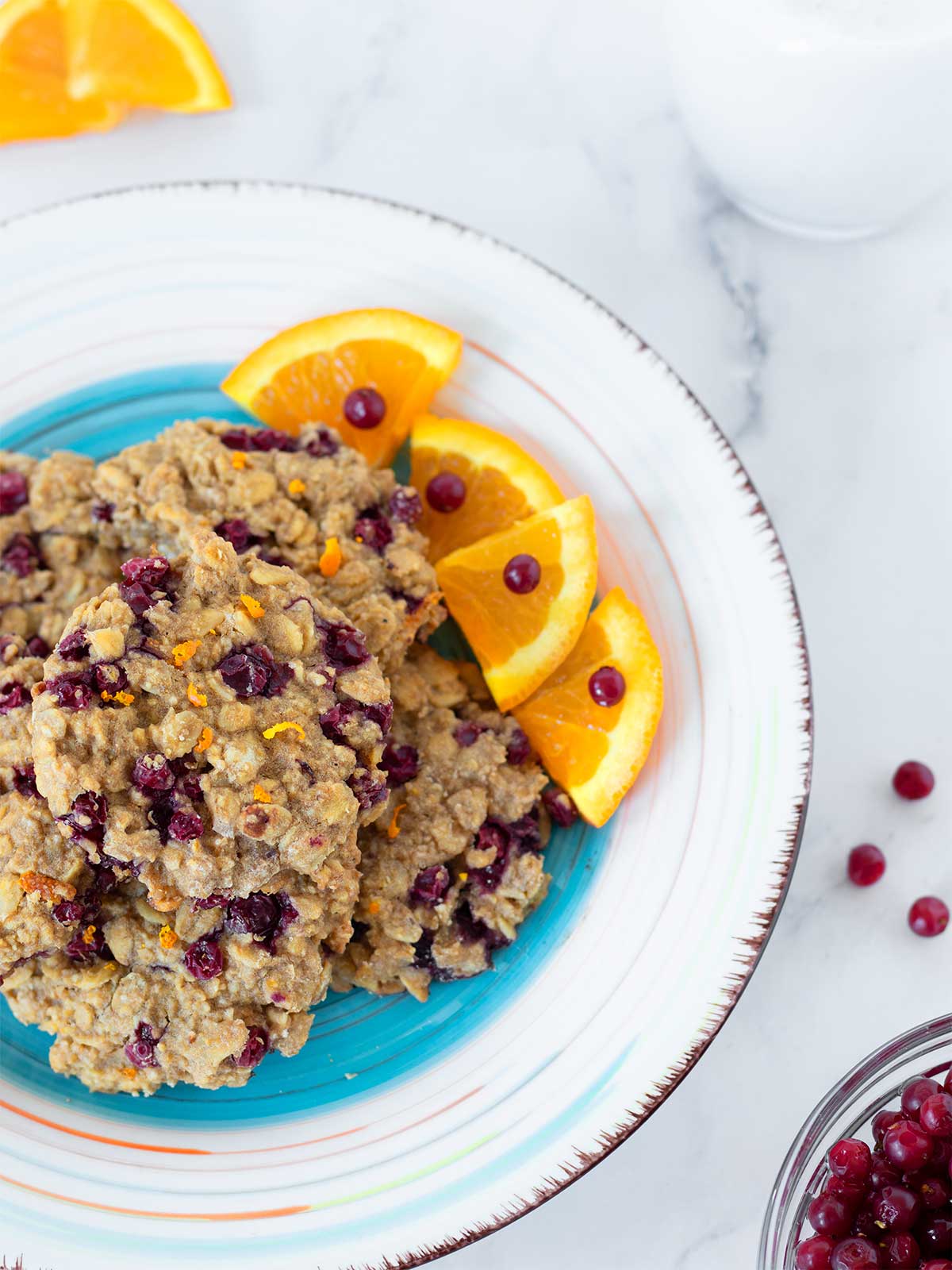 Chewy cranberry oatmeal cookies in a plate with sliced fresh orange and wild cranberries with a glass of dairy-free milk