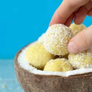 Vegan lemon coconut bliss balls in a coconut shell with frmale fingers holding one energy bite with blue background