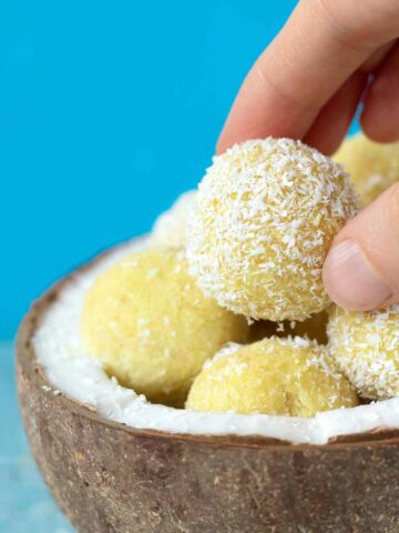 Vegan lemon coconut bliss balls in a coconut shell with frmale fingers holding one energy bite with blue background