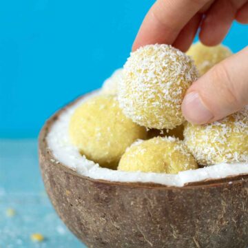 Vegan lemon coconut bliss balls in a coconut shell with frmale fingers holding one energy bite with blue background