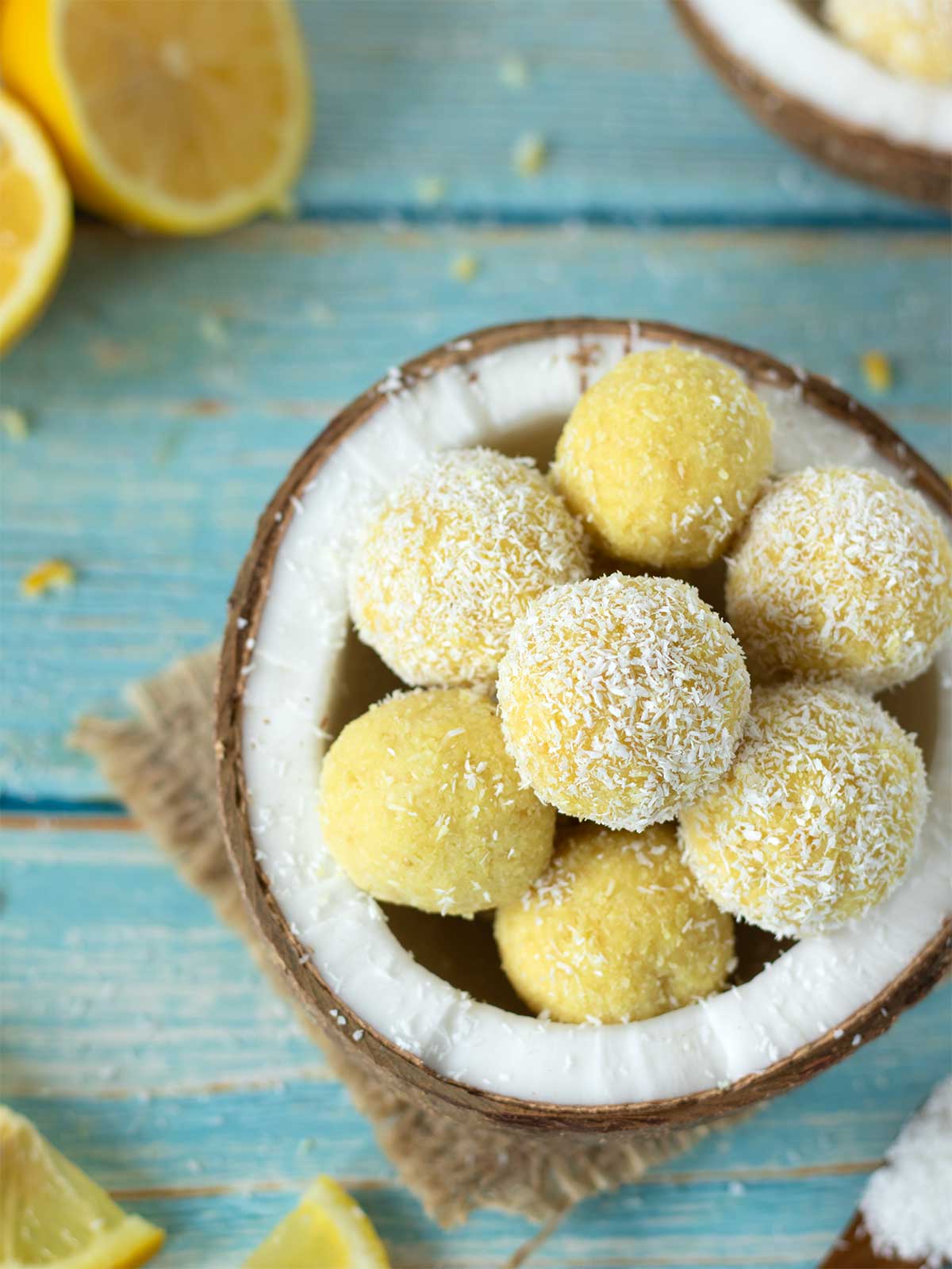 Lemon coconut balls in a coconut shell on blue wooden table with desiccated coconut flakes and sliced lemon.