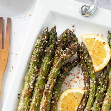 Vegan oven-roasted asparagus on white plate sprinkled with red pepper flakes and drizzled freshly squeezed lemon juice and wooden fork on white table