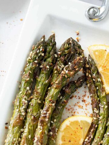 Vegan oven-roasted asparagus on white plate sprinkled with red pepper flakes and drizzled freshly squeezed lemon juice and wooden fork on white table