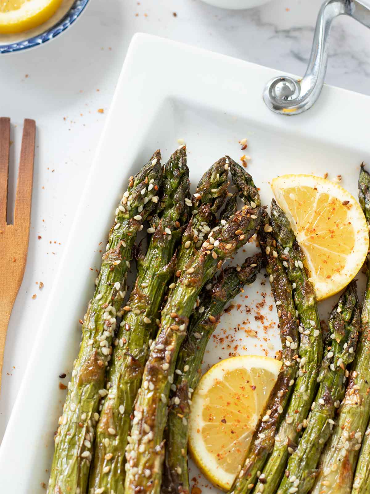 Healthy vegan oven-roasted asparagus on white plate sprinkled with red pepper flakes and drizzled freshly squeezed lemon juice and wooden fork on white table