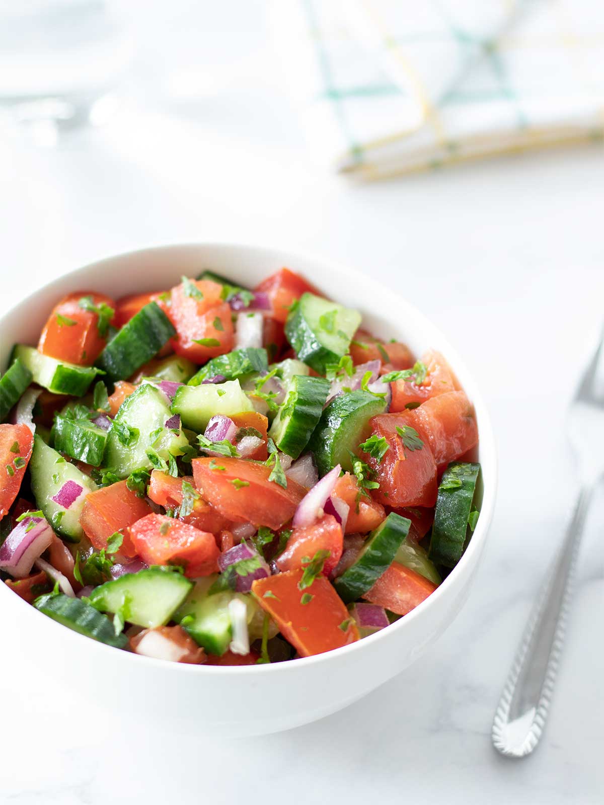 Cucumber and tomato salad with red onion and parsley in a bowl with fork on white marble table