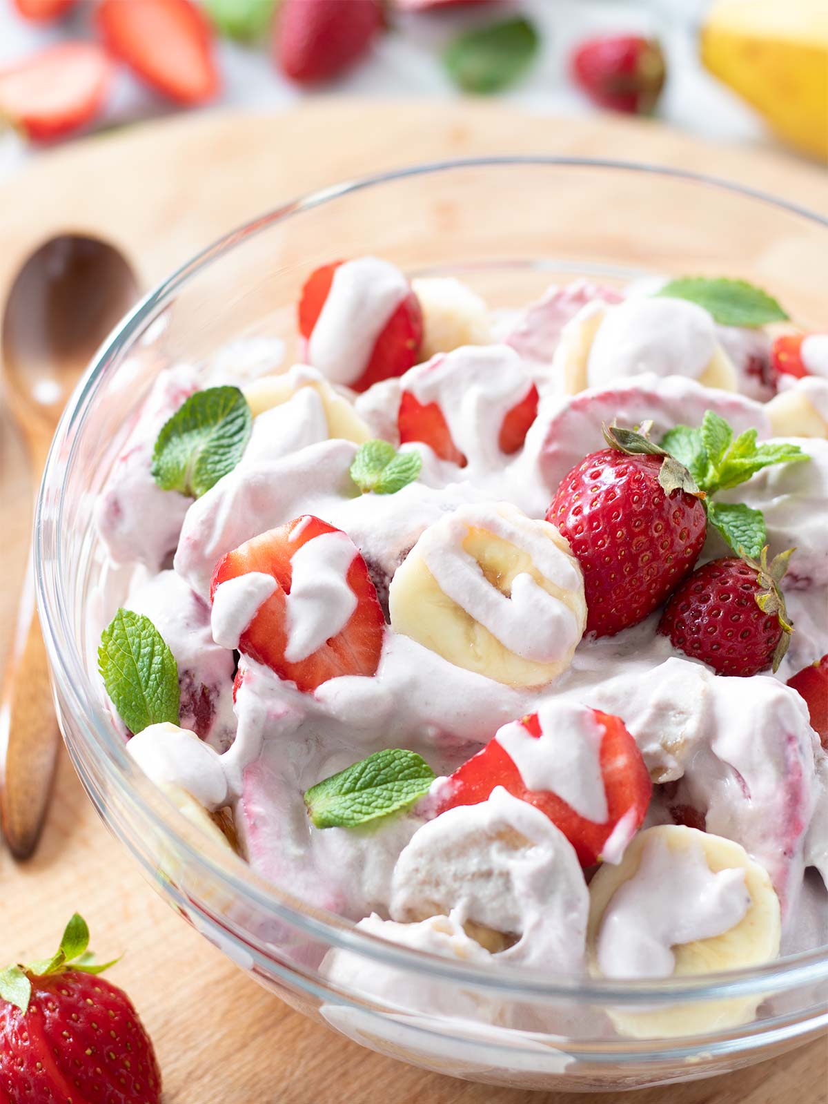 Dessert salad with creamy cheesecake dressing in a bowl with wooden spoon and fresh strawberries and banana in the background