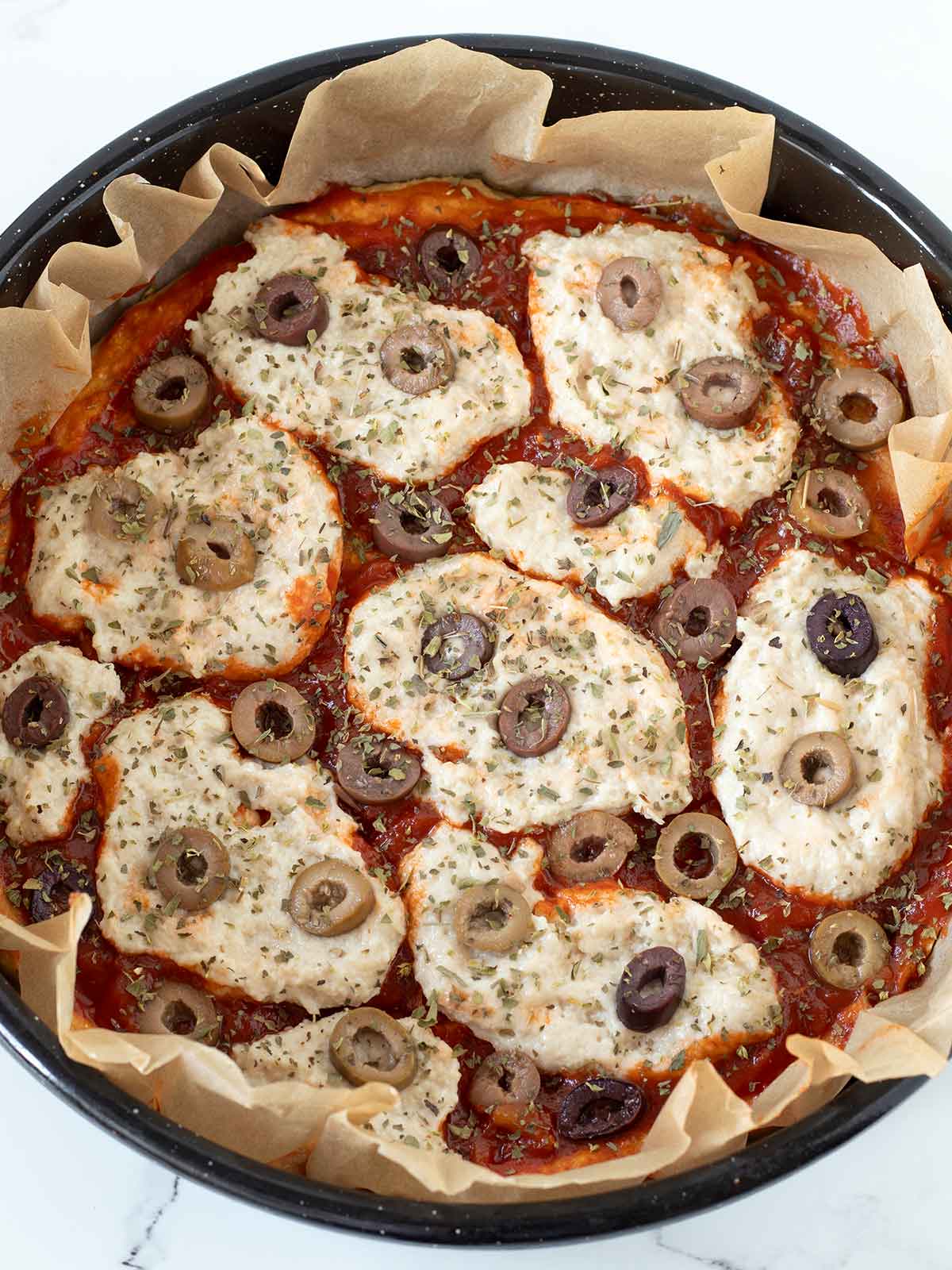 Chickpea flour pizza crust topped with vegan mozzarella cheese, tomato sauce and olives prepared for baking.